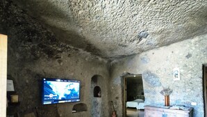 A rustic room with a television mounted on the wall, a wooden bench, a small table with a vase, and a bed in the background. - Las Margaritas Cave House (Artenara)
