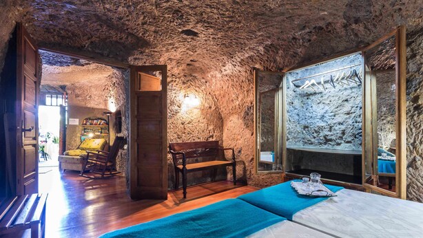 A rustic room with a wooden bench, a bed with a blue blanket, and a stone wall with a framed picture. - Las Margaritas Cave House (Artenara)