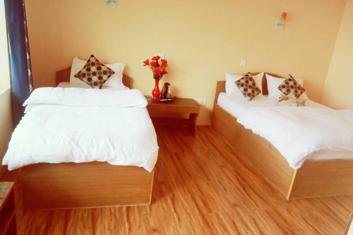 A hotel room with two wooden beds, each with white bedding and patterned pillows. A small table with a vase of red flowers is between the beds.