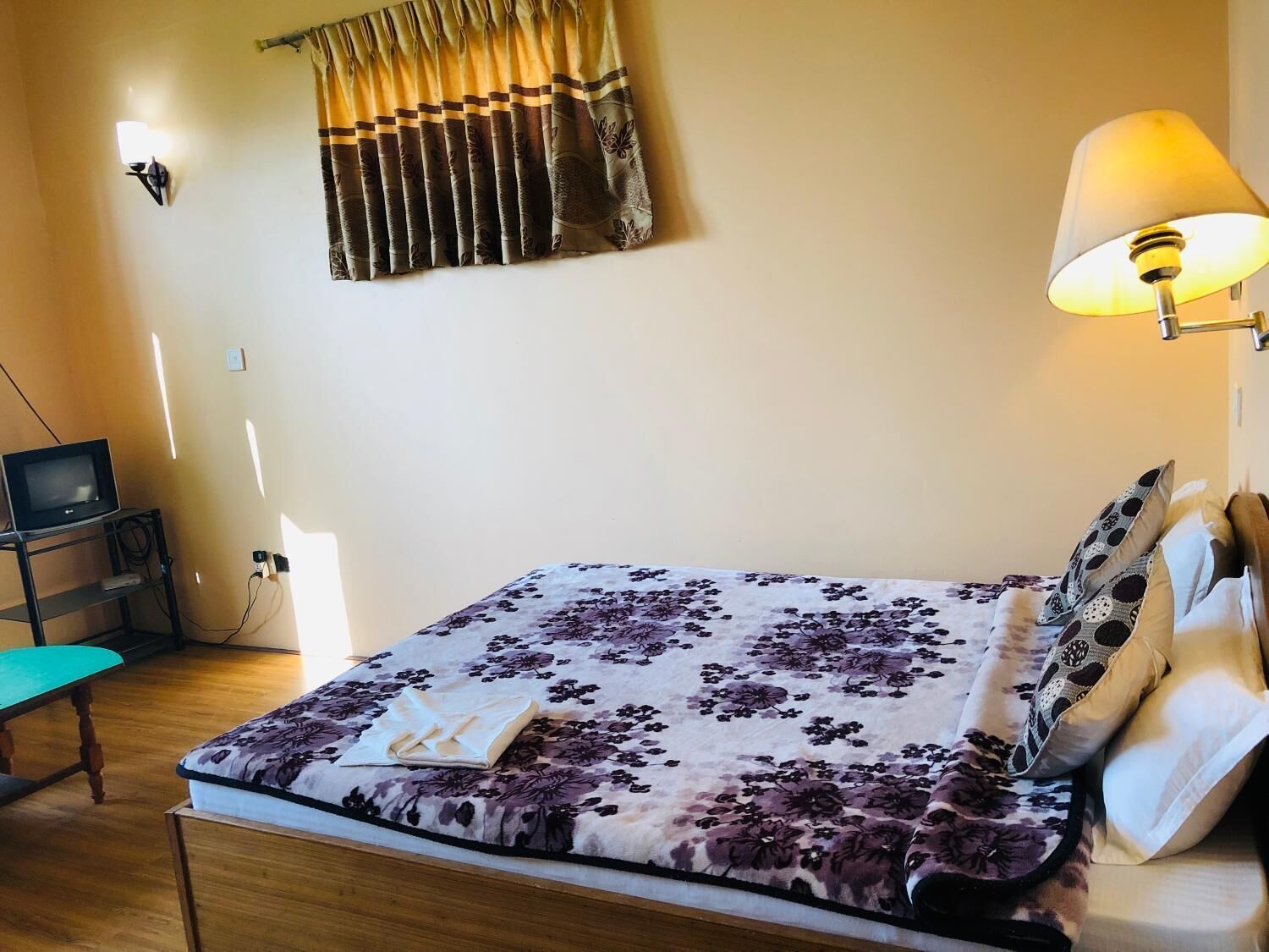 A neatly arranged bedroom with a bed, a bedside lamp, a television on a stand, and a window with curtains.