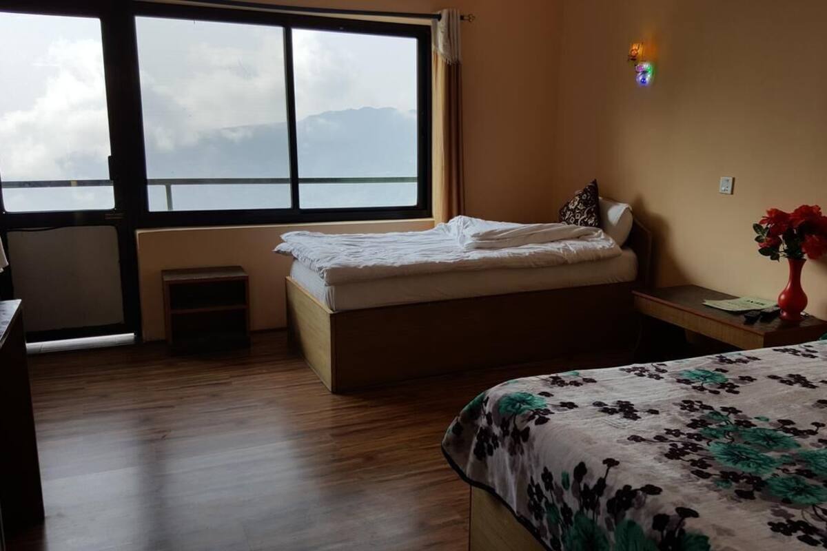 A bedroom with a bed, a window, a bedside table with a vase, and a view of mountains.