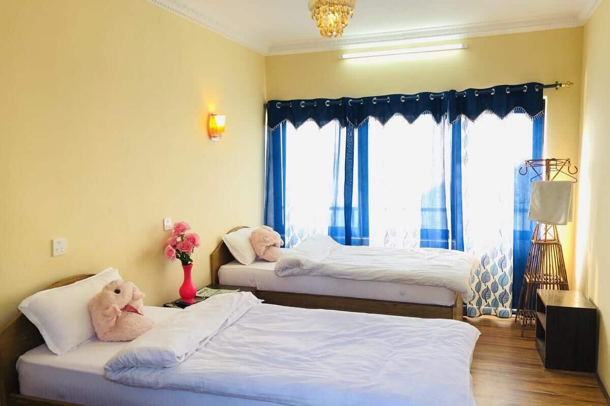 A hotel room with two beds, a bedside lamp, a vase with flowers, and a window with blue curtains.