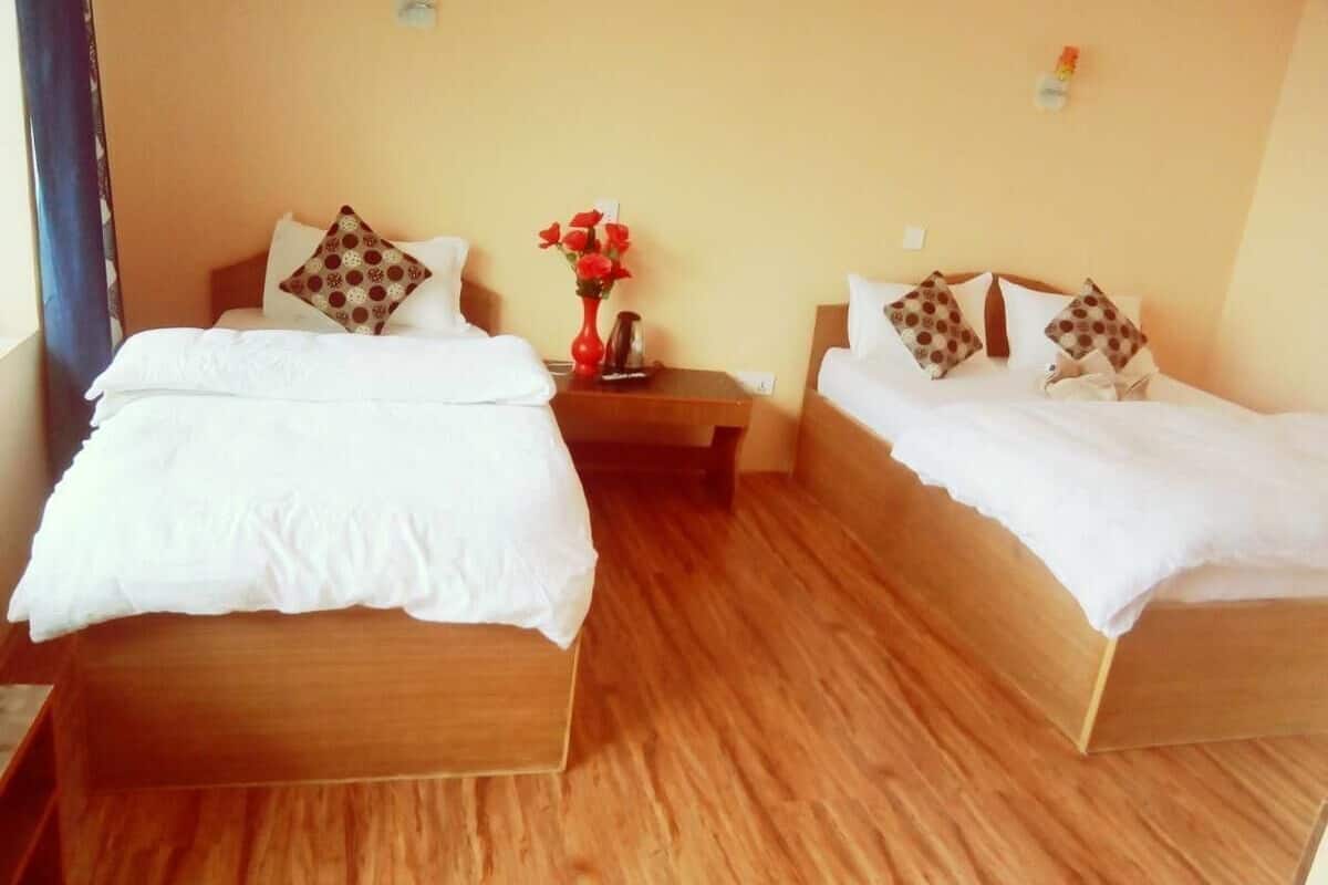 A room with two wooden beds, each with white bedding and patterned pillows, a small bedside table with a vase of red flowers, and a wooden floor.
