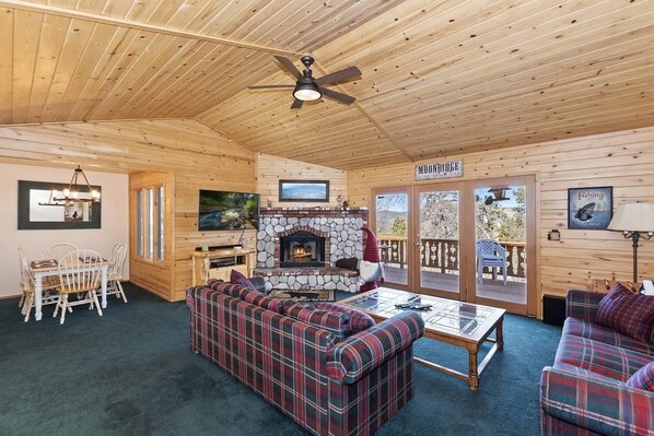 A cozy cabin interior with a stone fireplace, plaid sofas, a dining area, and a balcony.