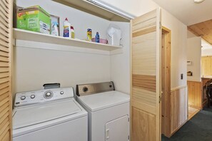 Een wasserette met twee wasmachines, een plank met schoonmaakmiddelen en een houten deur.