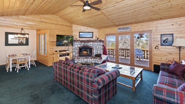 Ein gemütliches Blockhaus-Interieur mit Kamin aus Stein, Karomustern-Sofas, einem Essbereich und einer Terrasse mit dem Schild "MOONROSE".