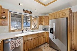 Een keuken in landhuisstijl met houten kasten, een koelkast van roestvrij staal en een raam met uitzicht op een besneeerd landschap.