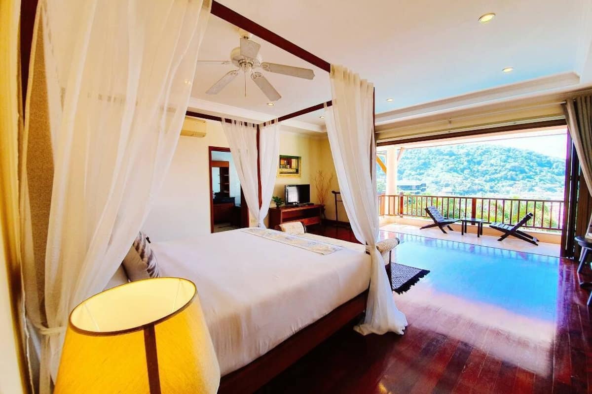 A four-poster bed with a canopy, a balcony with lounge chairs, and a view of a mountainous landscape.