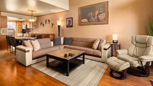 Una sala de estar con un sofá modular, una mesa de centro, un sillón reclinable, un comedor y una cocina al fondo, además de un cuadro en la pared.