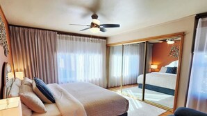 Habitación de hotel con cama, ventilador de techo, espejo grande, zona de estar y puerta corrediza que da a un balcón.