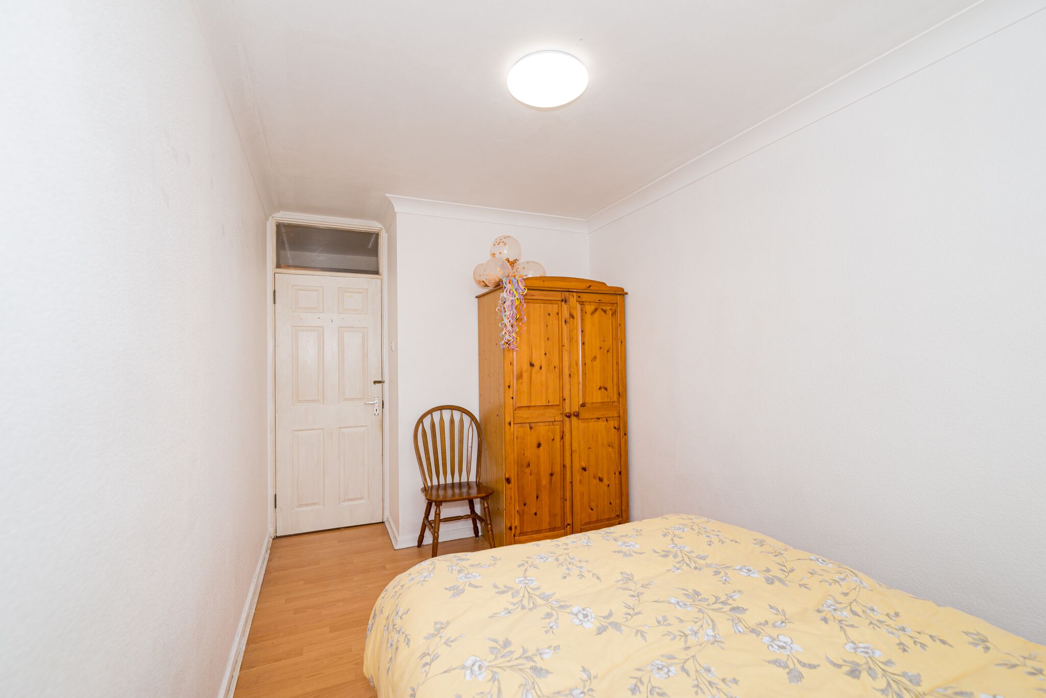 A bedroom with a wooden wardrobe, a floral bedspread, a wooden chair, and a white door.