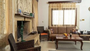 A living room with a fireplace, wicker furniture, a sofa with cushions, a coffee table with fruit, and a window with curtains. - Modern holiday home in Stalos with balcony (Stalos)