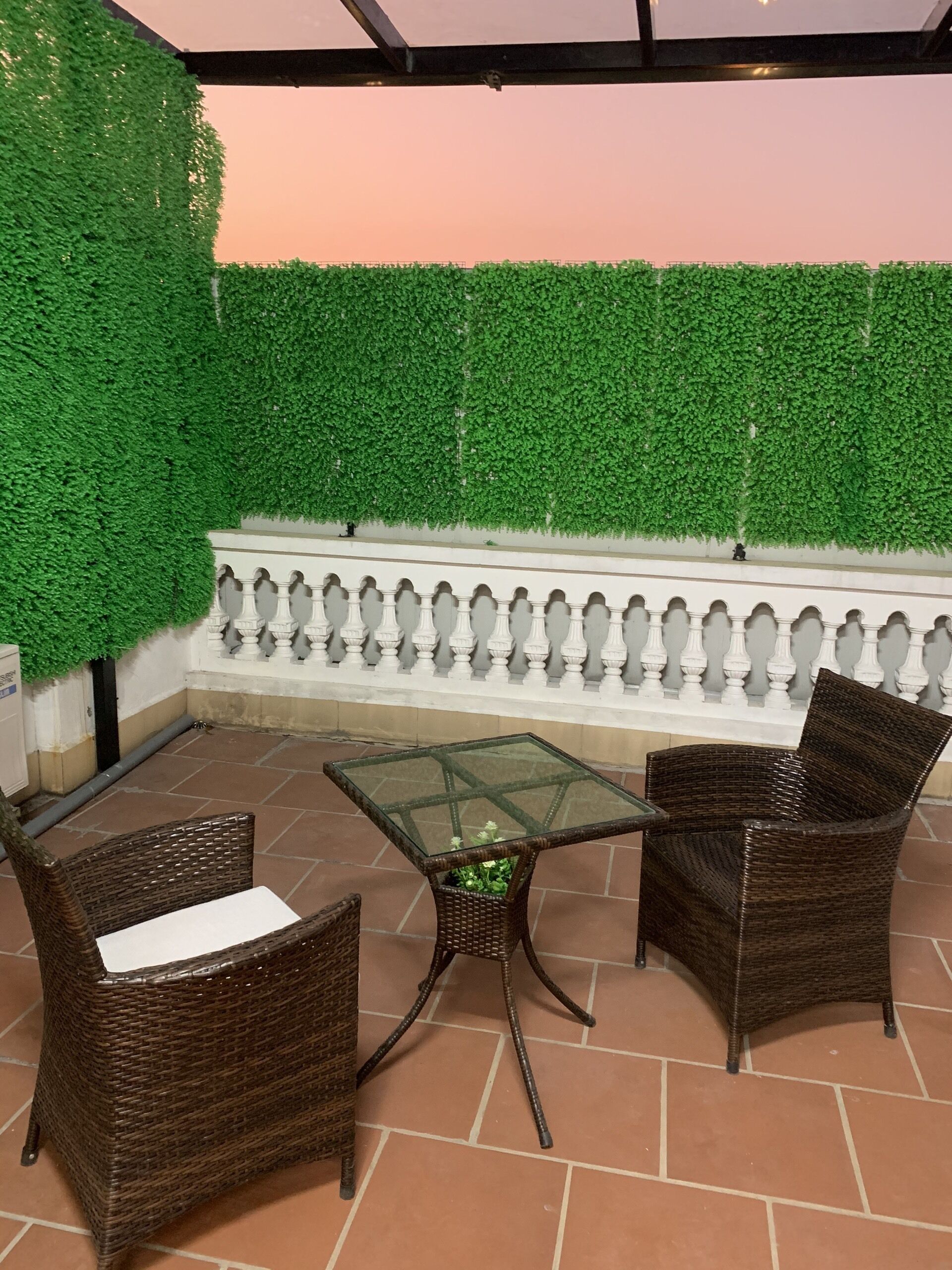 An outdoor patio with wicker furniture, a glass-top table, and a green artificial hedge wall.