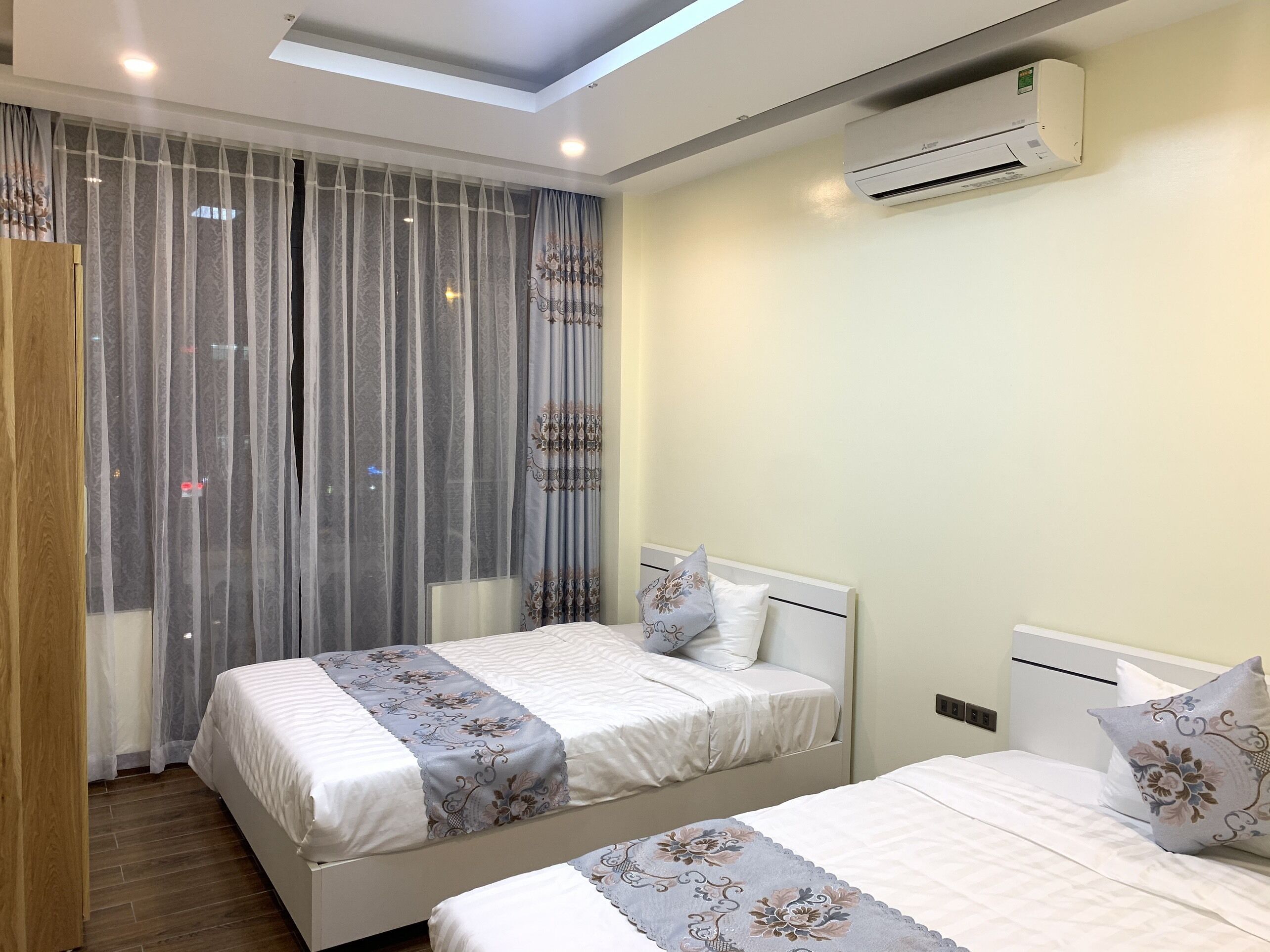 A hotel room with two beds, a skylight, an air conditioning unit, and a curtain with a floral pattern.