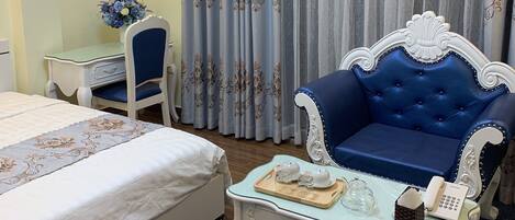 Una habitación de hotel con una cama, un sofá tapizado azul, una pequeña mesa blanca con un teléfono y una bandeja de objetos, y una silla.