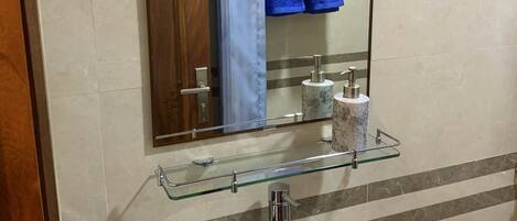 Un baño con un lavamanos blanco, estante de vidrio con artículos de aseo, espejo y toallero con toallas azules.