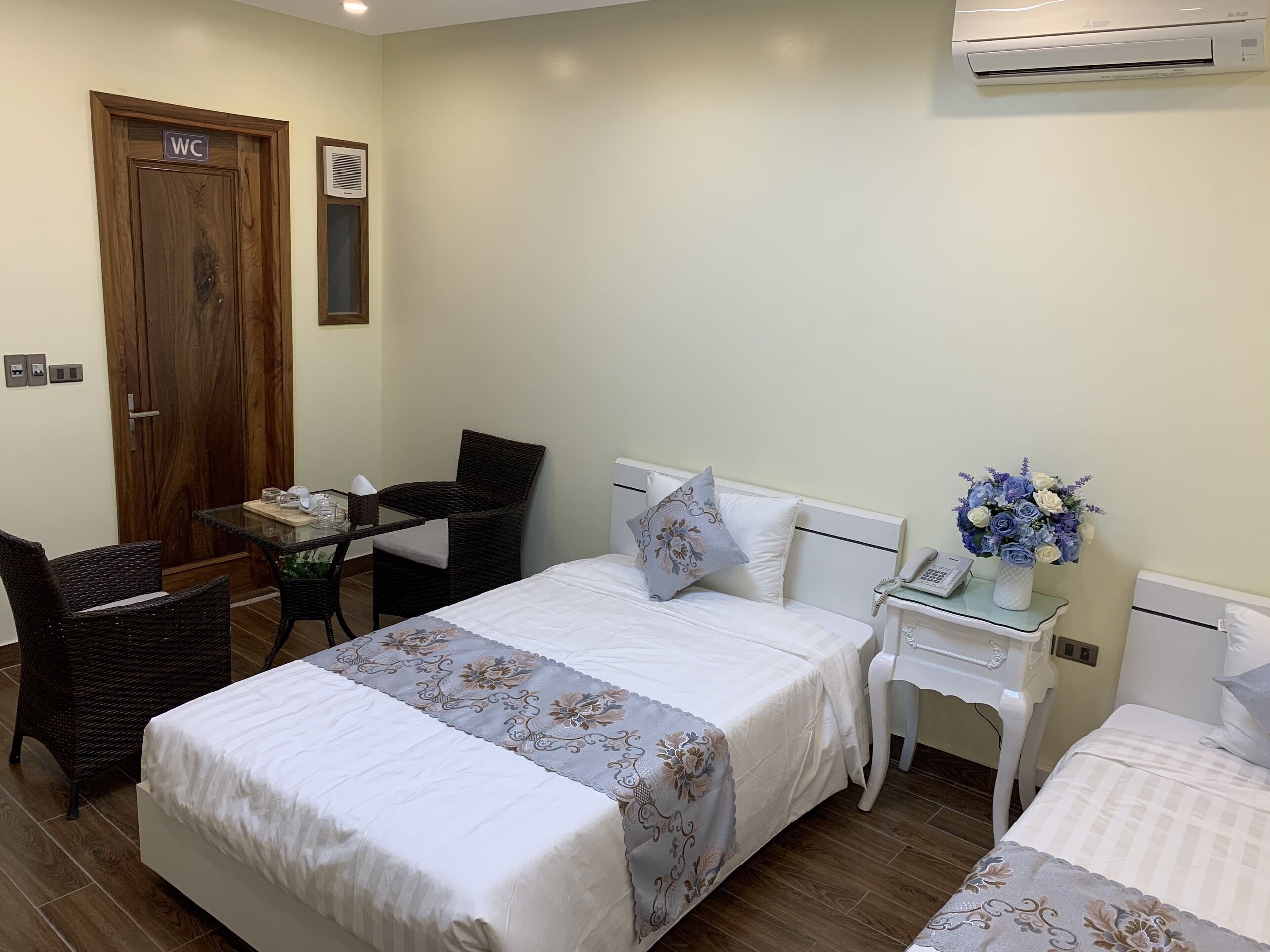 A hotel room with a bed, a small table with chairs, a bedside table with a phone and vase, and an air conditioning unit on the wall.