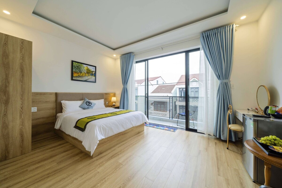 A hotel room with a bed, a desk with a mirror, a chair, a balcony with a view, and a window with curtains.