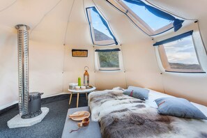 Interior acogedor de una tienda de campaña con una cama, un calentador, una mesita con objetos y ventanas que ofrecen vistas del exterior.