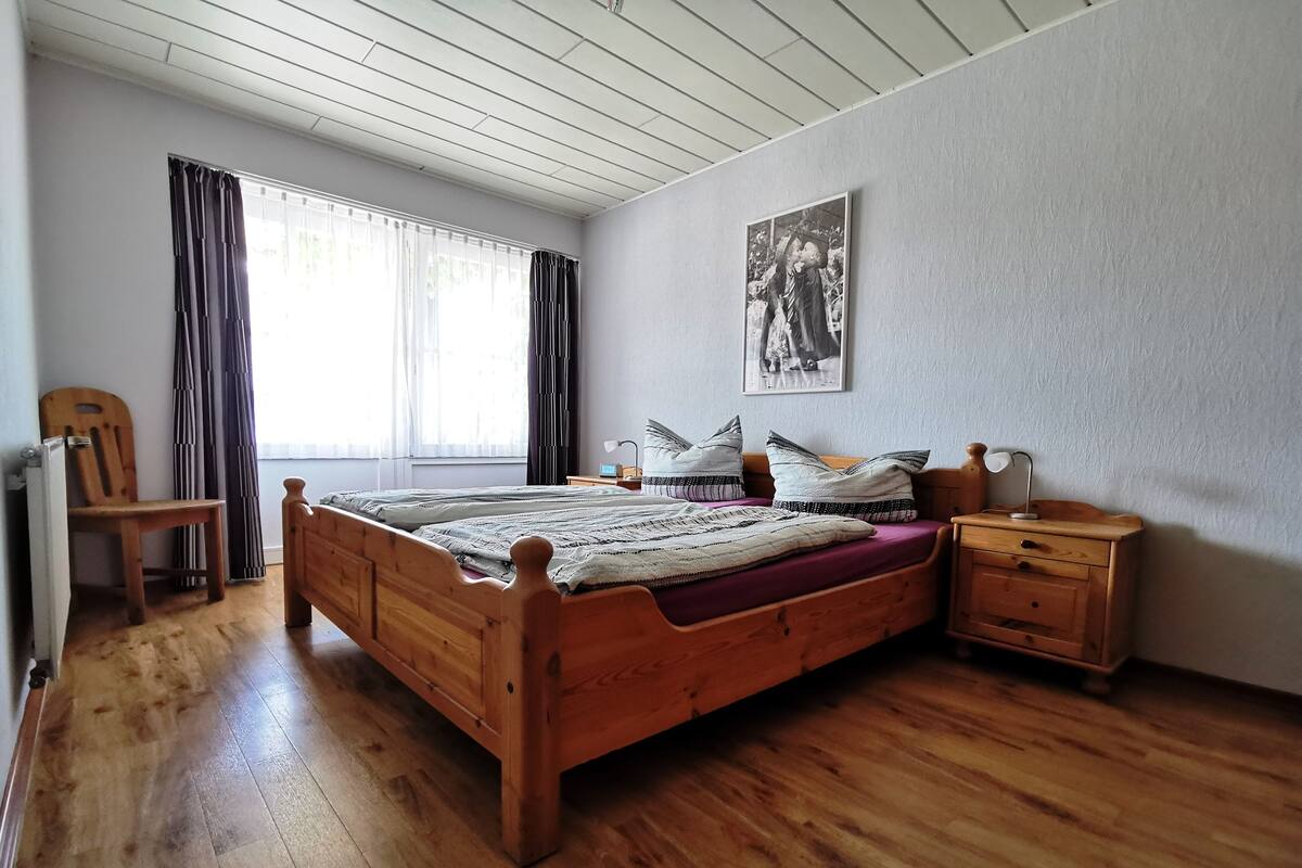 Ein Schlafzimmer mit einem hölzernen Bett, einem Sessel, einem Nachttisch und einem Fenster mit Vorhängen.