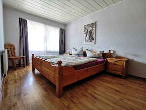 A bedroom with a wooden bed, a chair, a nightstand, and a window with curtains.