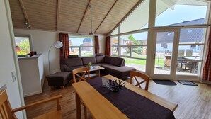 Un salon chaleureux avec un plafond en bois, un canapé, une table à manger et de grandes fenêtres donnant sur l’extérieur.