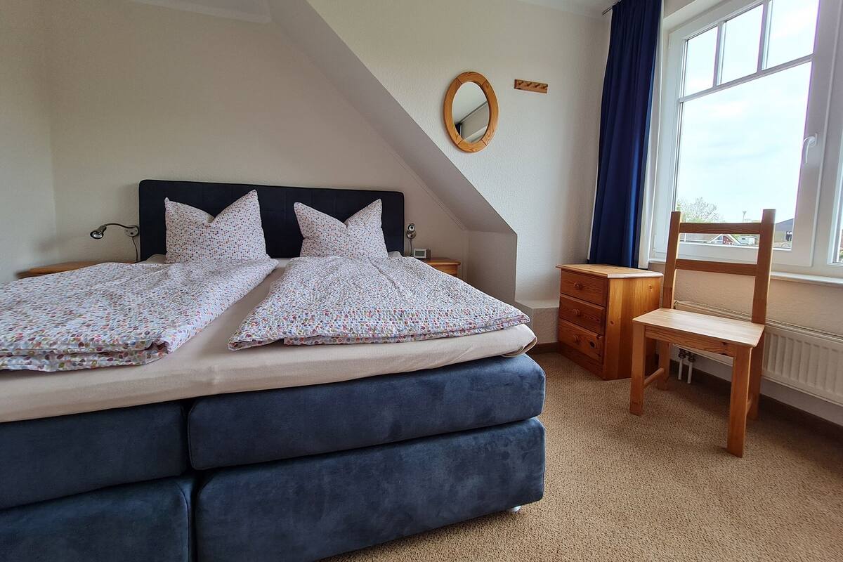 Ein Schlafzimmer mit Bett, Holzstuhl, Nachttisch, Spiegel und Fenster mit Vorhängen.