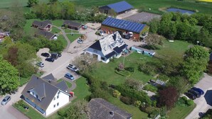 Vue aérienne d’une propriété rurale comprenant plusieurs bâtiments, notamment une maison avec un toit en pente, une grange et une installation de panneaux solaires. Plusieurs voitures sont garées et la propriété est entourée de champs verdoyants et d’une étendue d’eau.