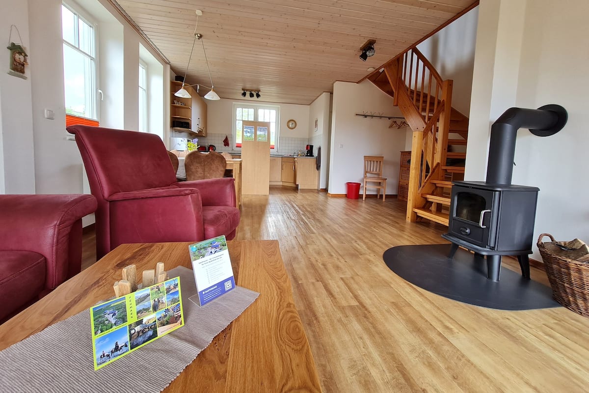 Ein gemütliches Wohnzimmer mit einem roten Sofa, Holzböden, einem schwarzen Kaminofen und einer Küche im Hintergrund.