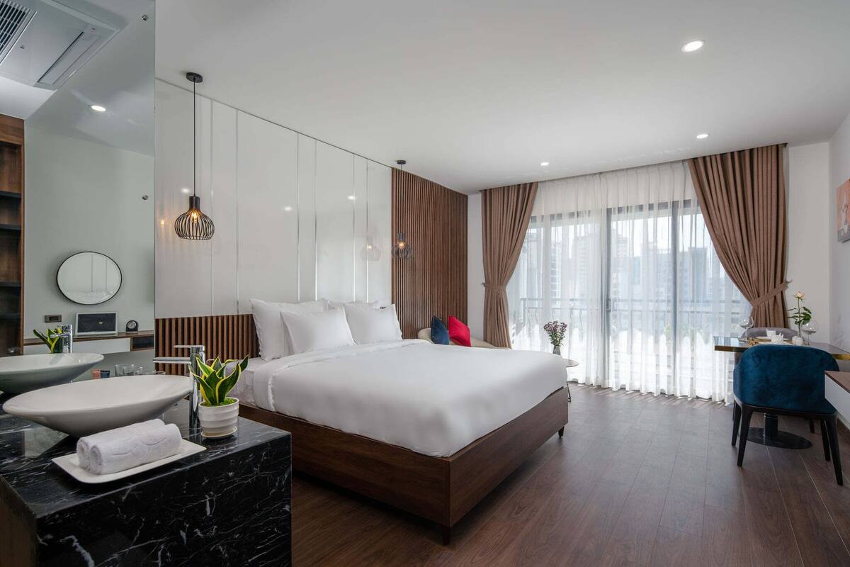 A modern hotel room with a large bed, a sitting area with a desk, a bathroom with a large sink, and a view of the city through large windows.