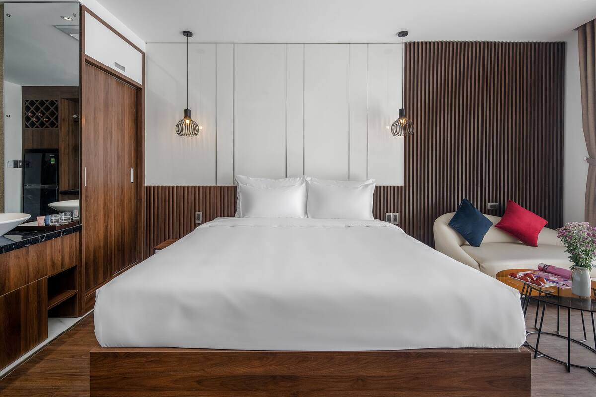 A modern hotel room with a large bed, wooden wardrobe, and a seating area with a sofa and armchairs.