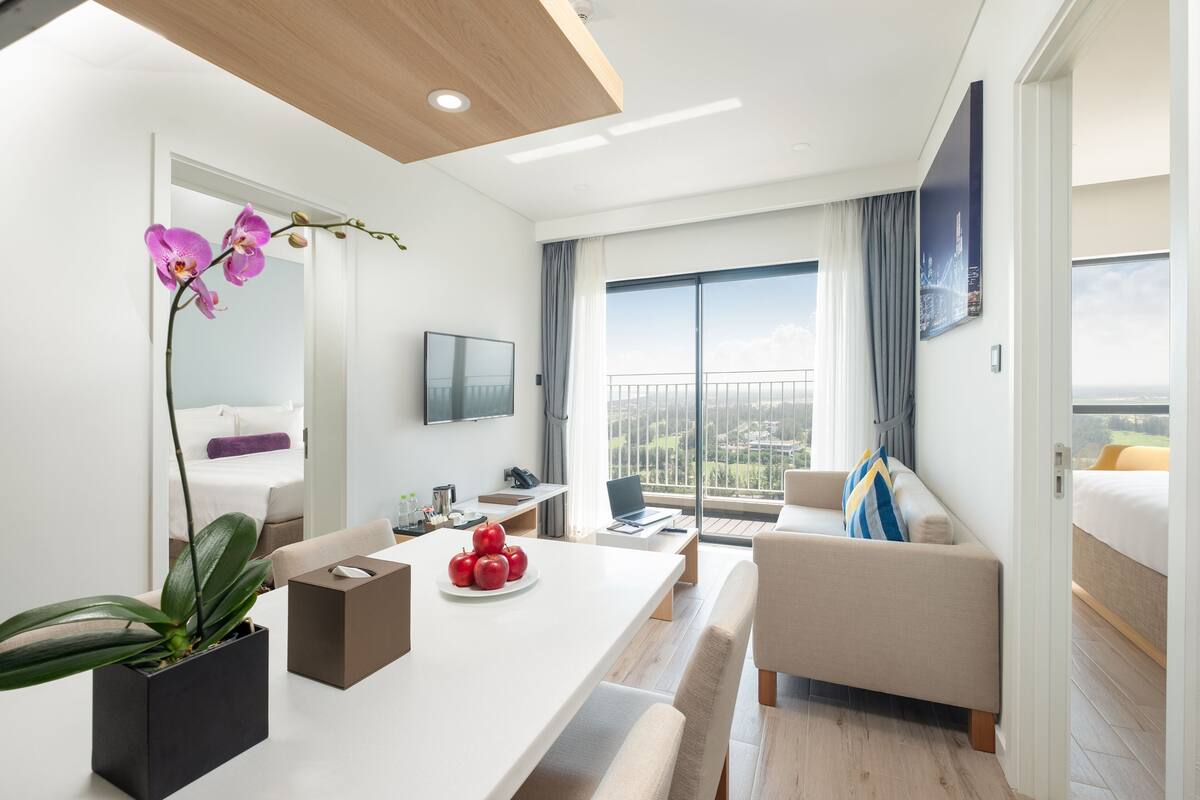 Une chambre d’hôtel moderne avec un balcon, un coin repas avec une table blanche, un canapé et une plante en pot.