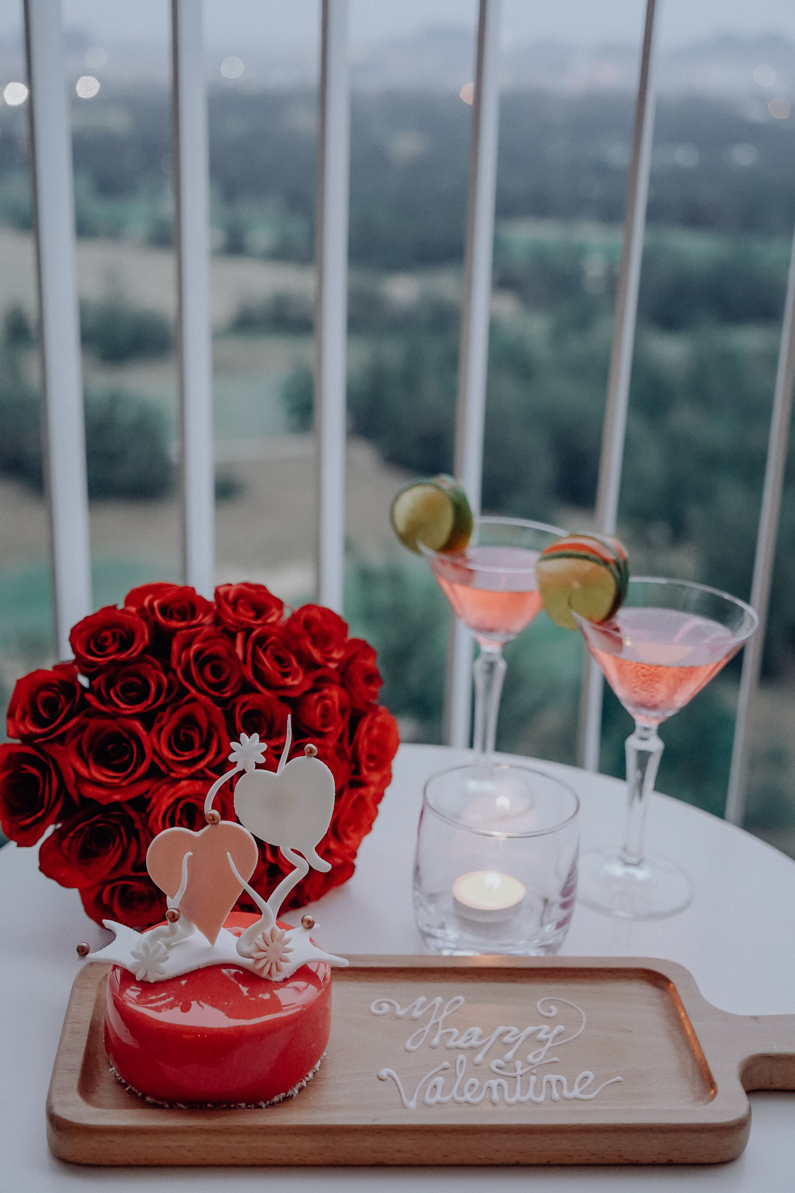 A red cake with "Happy Valentine's Day" on a wooden tray, a bouquet of red roses, and two martini glasses with lime slices.