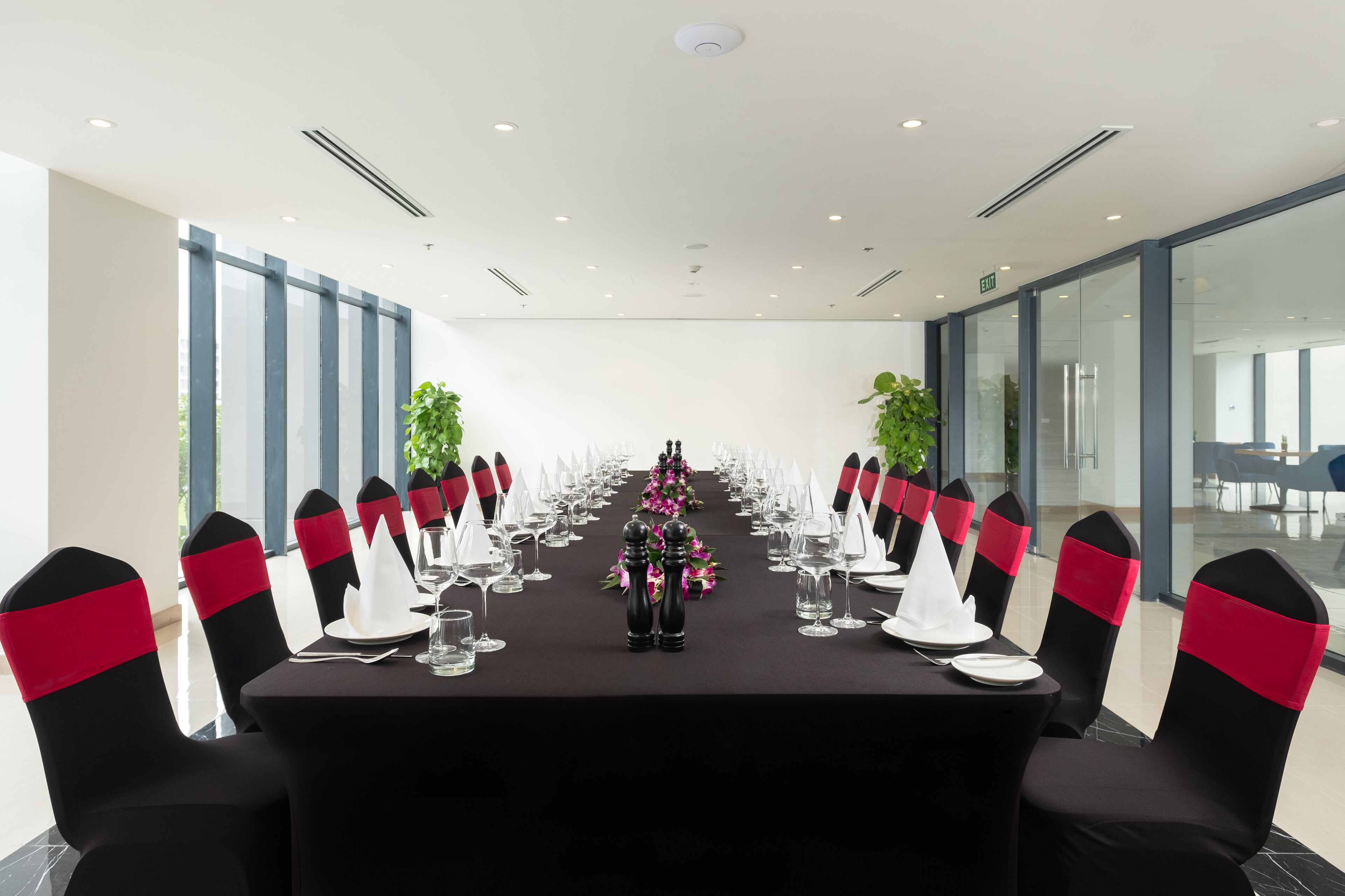 A conference room with a long table, black and red sashed chairs, and a floral centerpiece.
