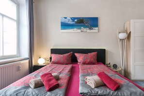 A double bed with pink and grey bedding, a painting of a tree on the wall, and a bedside table with a lamp.
