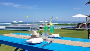 Une table à manger est dressée avec une bouteille, des verres et une bougie, et offre une vue sur une piscine et une plage, avec des parasols et des chaises longues.