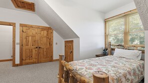 A bedroom with a sloped ceiling, wooden wardrobe, a bed with floral bedding, and a window with blinds. - Views, privacy, Kids Playhouse, Easy Access, Fast WiFi! Gated community (Waynesville)