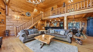 A spacious log cabin interior with a grand staircase, a cozy living area with two sofas, a dining table, and a kitchen area. - Views, privacy, Kids Playhouse, Easy Access, Fast WiFi! Gated community (Waynesville)