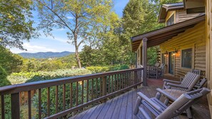 A wooden deck with a railing, a seating area with chairs, and a view of a forested landscape with mountains in the distance. - Views, privacy, Kids Playhouse, Easy Access, Fast WiFi! Gated community (Waynesville)