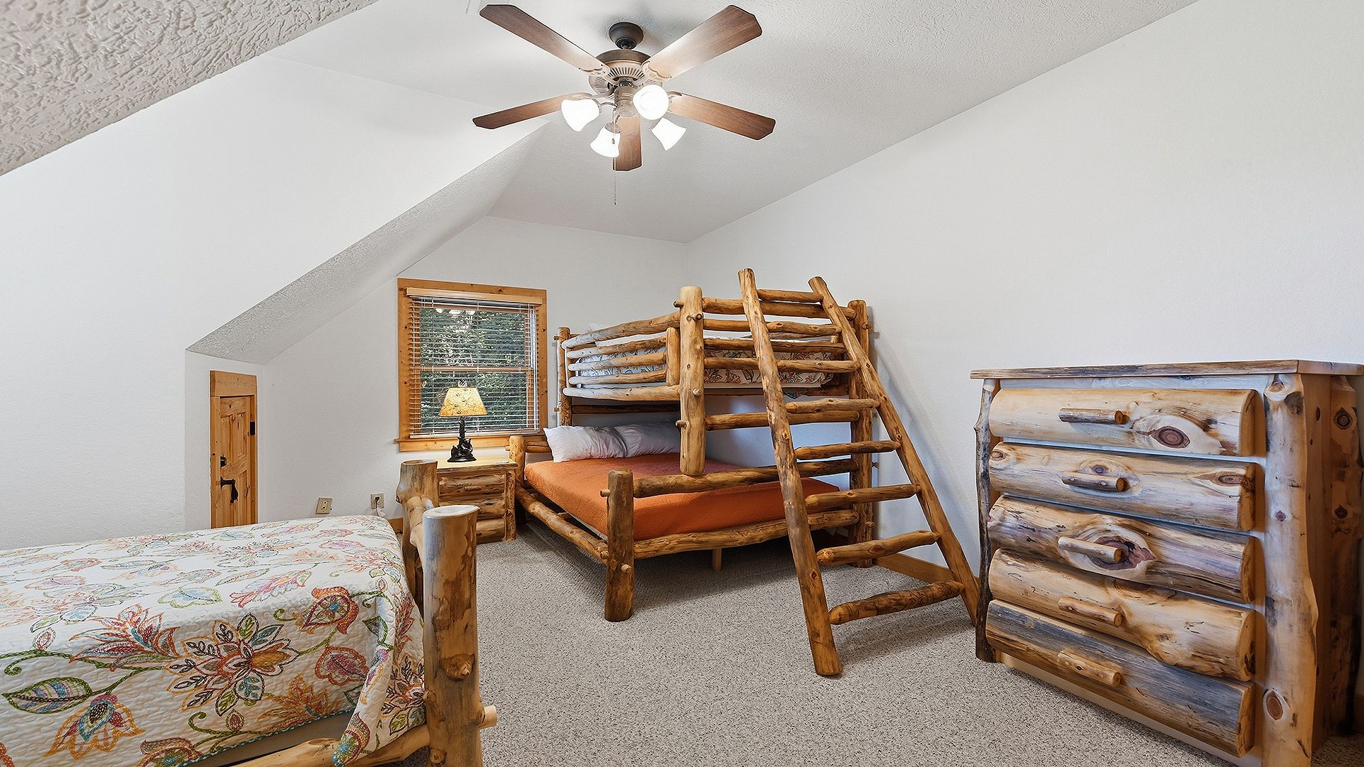 A bedroom with a sloped ceiling, a bed with a floral bedspread, a wooden ladder, a dresser, and a ceiling fan.