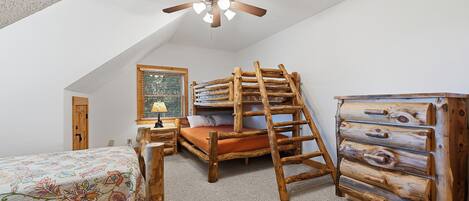 A bedroom with a sloped ceiling, a bed with a floral bedspread, a wooden ladder, a dresser, and a ceiling fan.