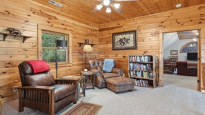 A cozy log cabin interior with a leather armchair, a cushioned sofa, a bookshelf, and a ceiling fan. - Views, privacy, Kids Playhouse, Easy Access, Fast WiFi! Gated community (Waynesville)