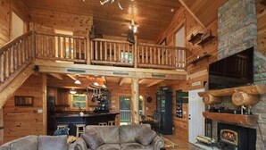 A spacious log cabin interior with a central seating area, a stone fireplace, and a balcony overlooking the yard. - Views, privacy, Kids Playhouse, Easy Access, Fast WiFi! Gated community (Waynesville)