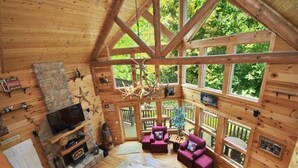 A log cabin interior with a stone fireplace, wooden furniture, and large windows offering a view of the outdoors. - Views, privacy, Kids Playhouse, Easy Access, Fast WiFi! Gated community (Waynesville)