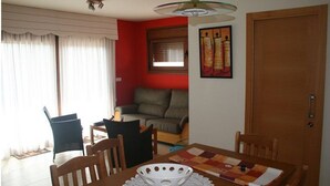 Een eetgedeelte met een houten tafel, stoelen en een decoratief middelpunt. Achtergrond is een bank en een raam met gordijnen.