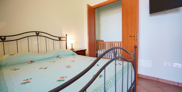 Un lit simple avec un cadre en métal et une couvre-lit à motifs floraux, une table de chevet avec une lampe, et une porte en bois donnant sur une autre pièce.
