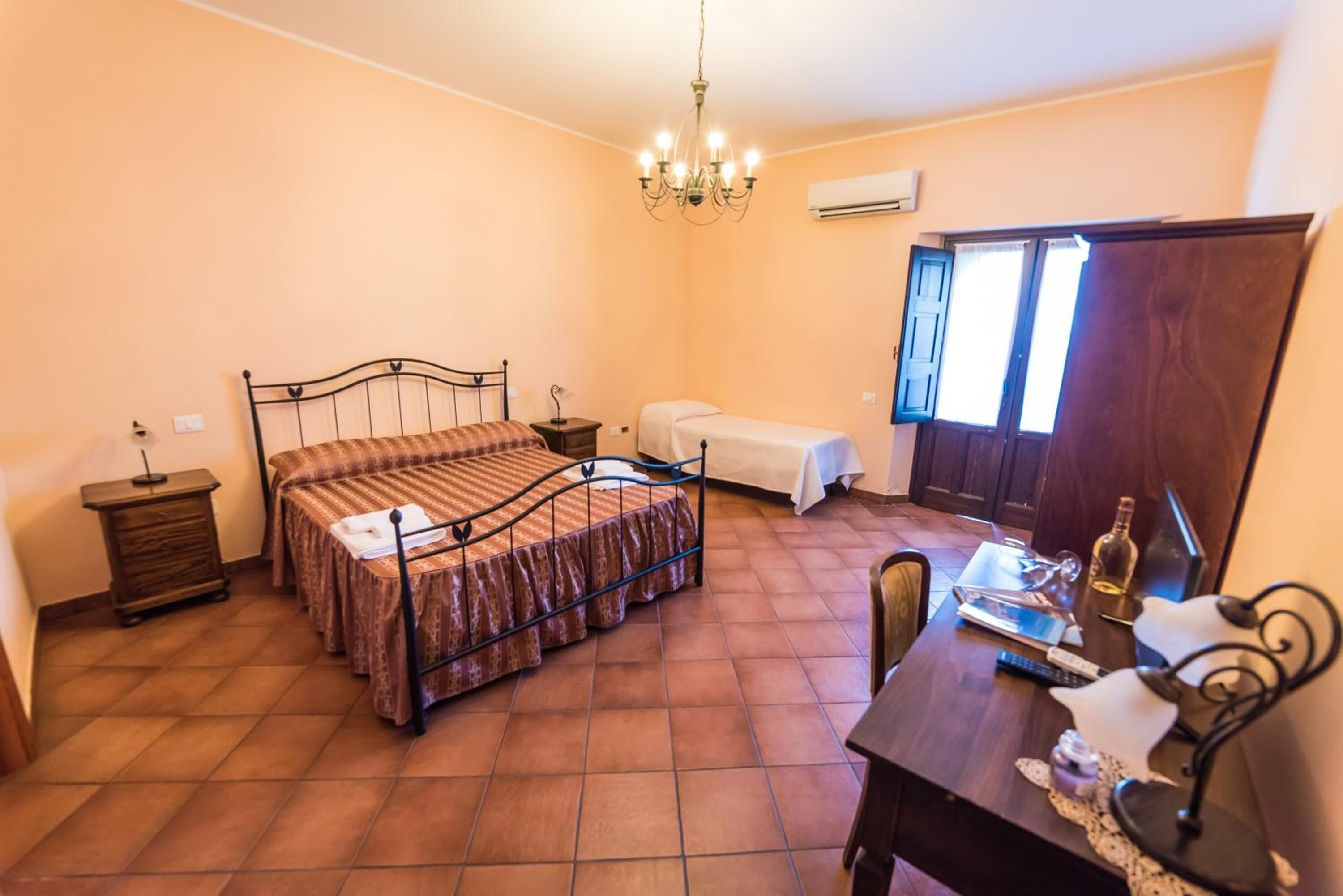 Un dormitorio con cama, mesitas de noche, un escritorio con silla, una araña y una ventana con cortinas.