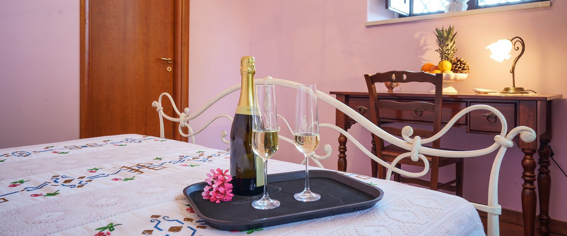 A bed with a decorative bedspread, a bedside table with a bottle of champagne and two glasses, and a wooden chair with a fruit bowl and a lamp.