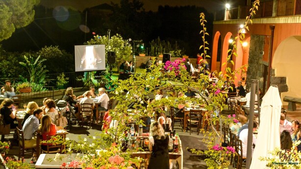 An outdoor dining area with tables and chairs, surrounded by plants and flowers.