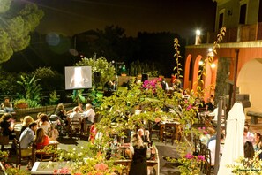 Un área de comedor al aire libre con mesas y sillas, rodeada de plantas y flores.